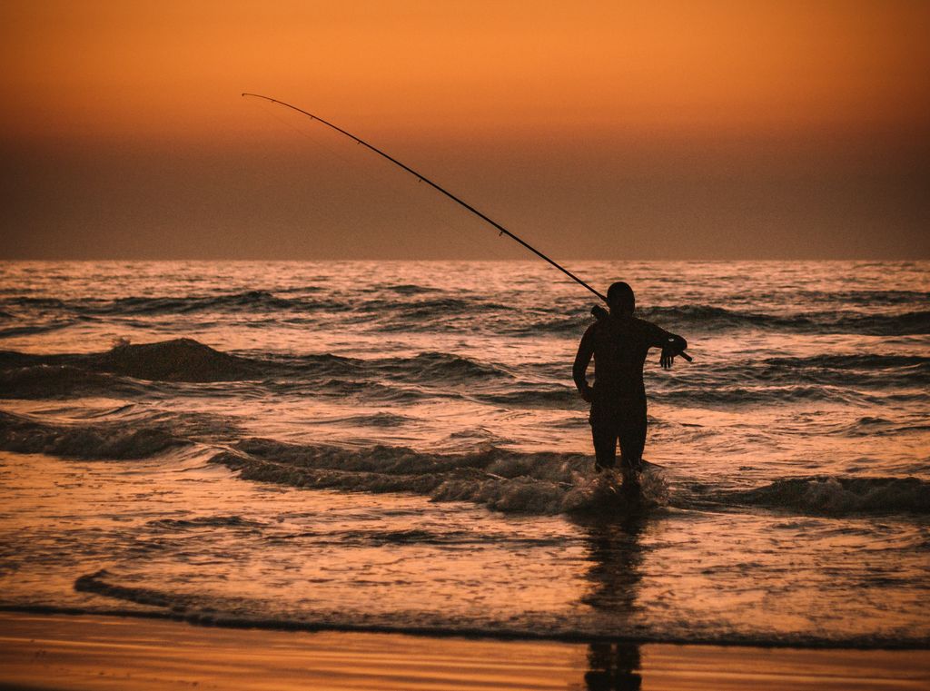 A dramatic silhouette of a fisherman standing in the sea at sunset, casting his line into the waves.