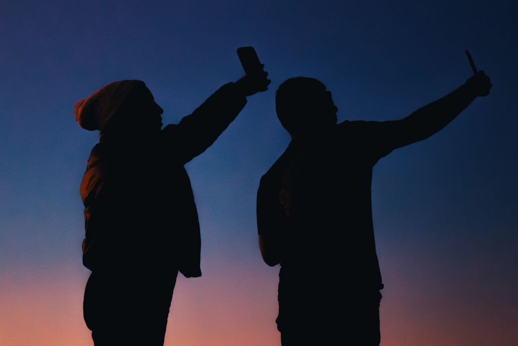 Silhouetted duo capturing selfies at twilight against a colorful sky.