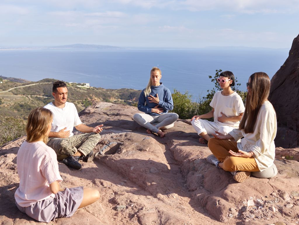 A diverse group of people meditating together on a rocky cliffside by the ocean.