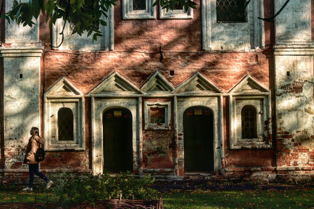 A historic monastery facade in Russia with sunlight casting shadows, a woman walking by.