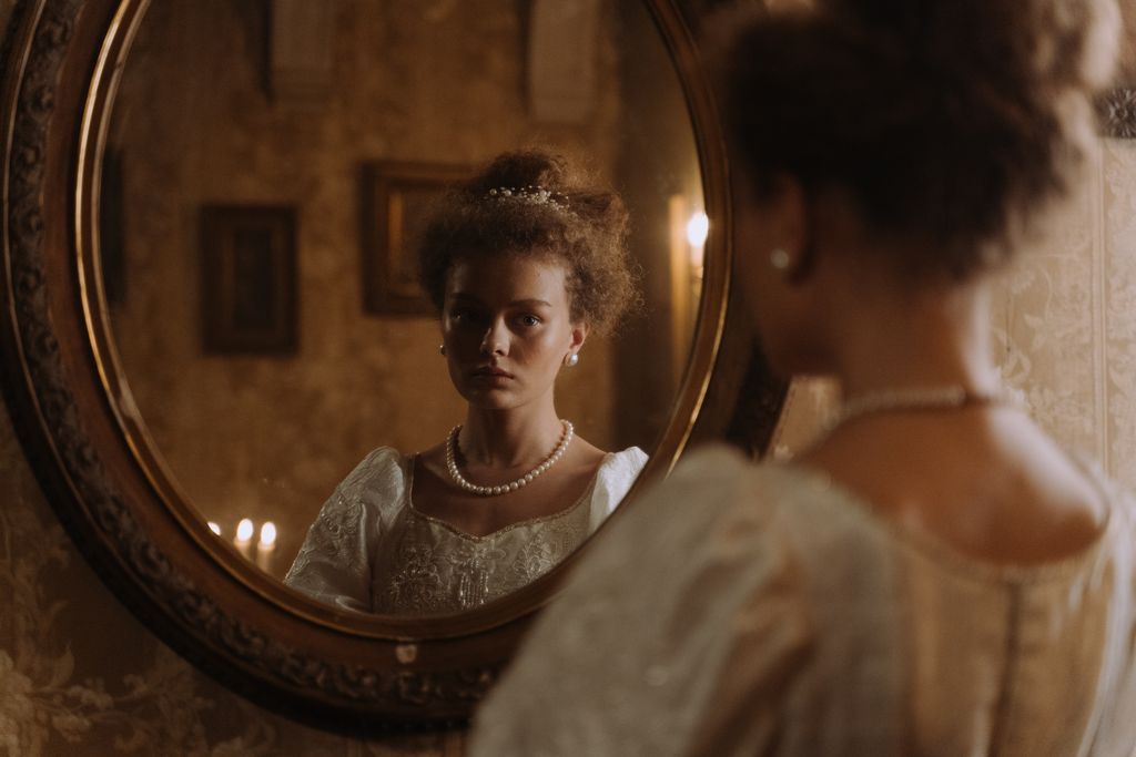A woman in a vintage empire style dress elegantly gazing at her reflection in an ornate mirror.