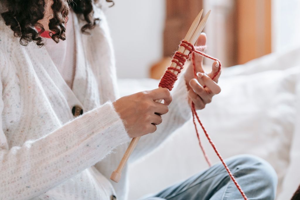 Crop unrecognizable female in casual wear knitting with needles while sitting on bed with cushion in light room at home