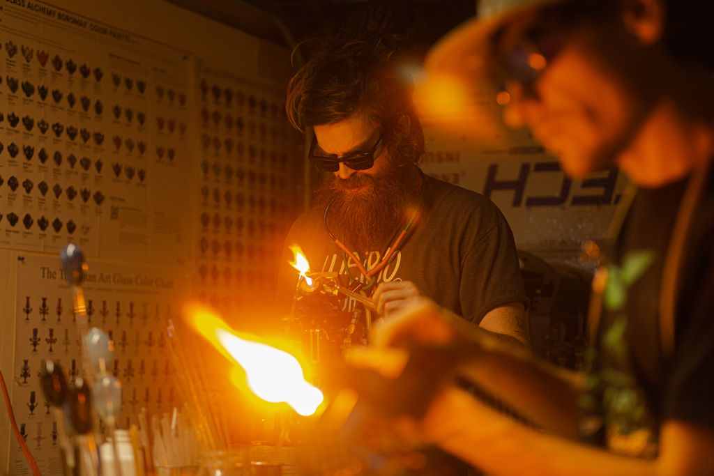 Two male artisans skillfully shape glass with fire in a dimly lit workshop.